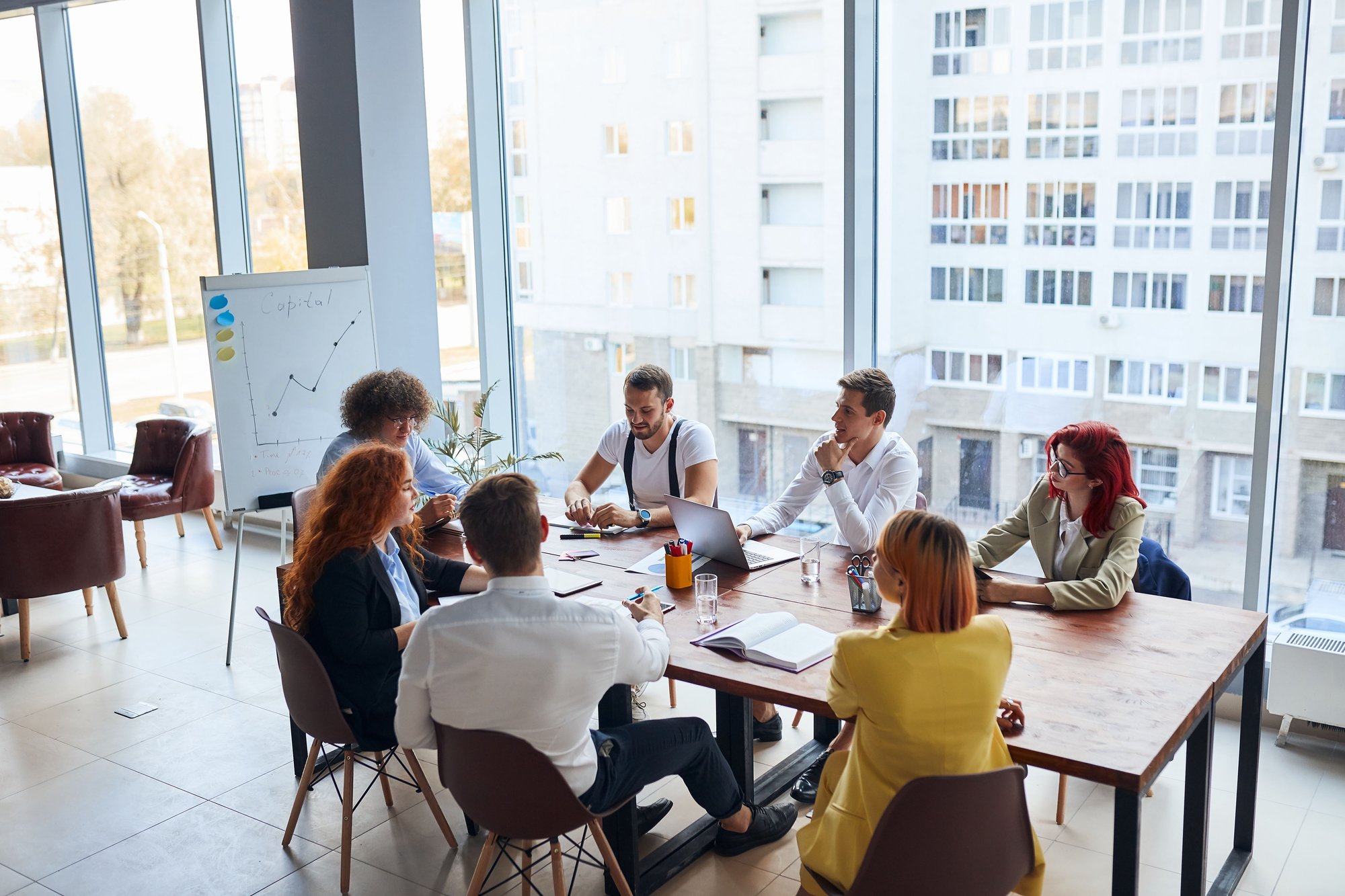 Young colleagues and business partners working as designers sit together at table in modern office with panoramic window. 